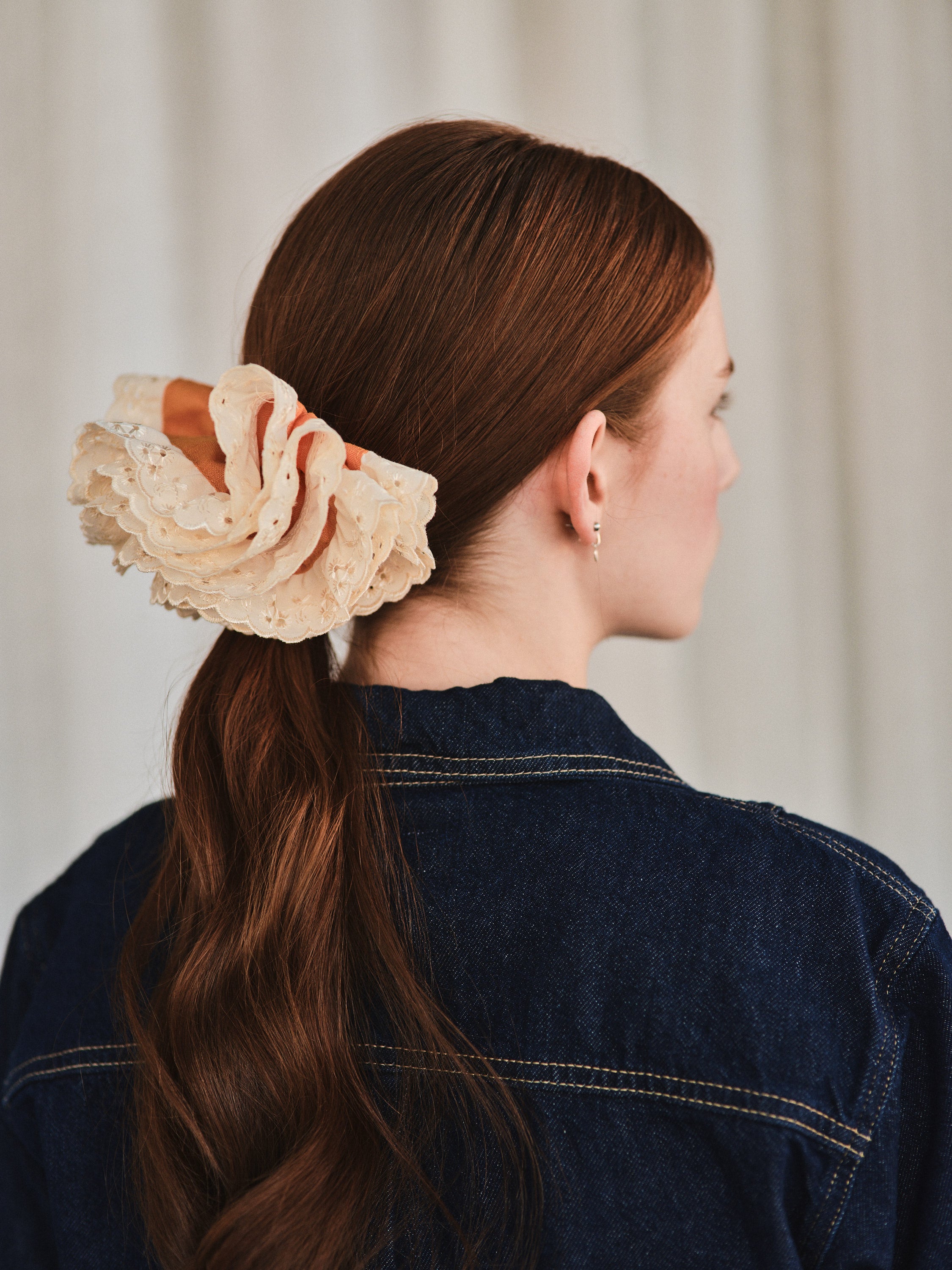 Azalea hair clip featuring guava orange linen and off-white eyelet lace, worn over a ponytail on a woman with natural long red hair. She’s wearing a dark denim shirt and is shown from a back profile with her head to the side.