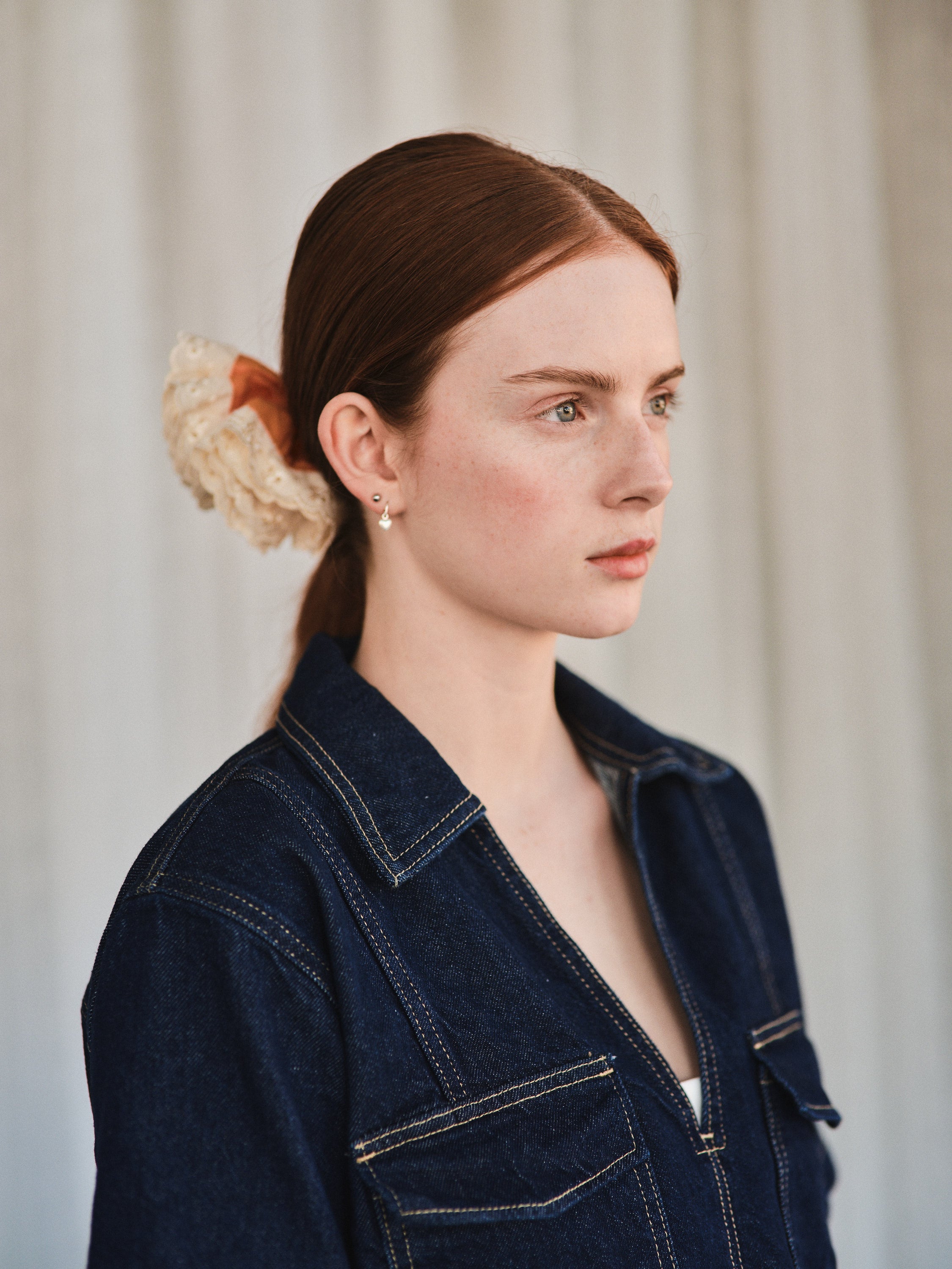 Azalea hair clip featuring guava orange linen and off-white eyelet lace, worn over a ponytail on a woman with natural long red hair. She’s wearing a dark denim shirt and is shown from a 3/4 front profile.
