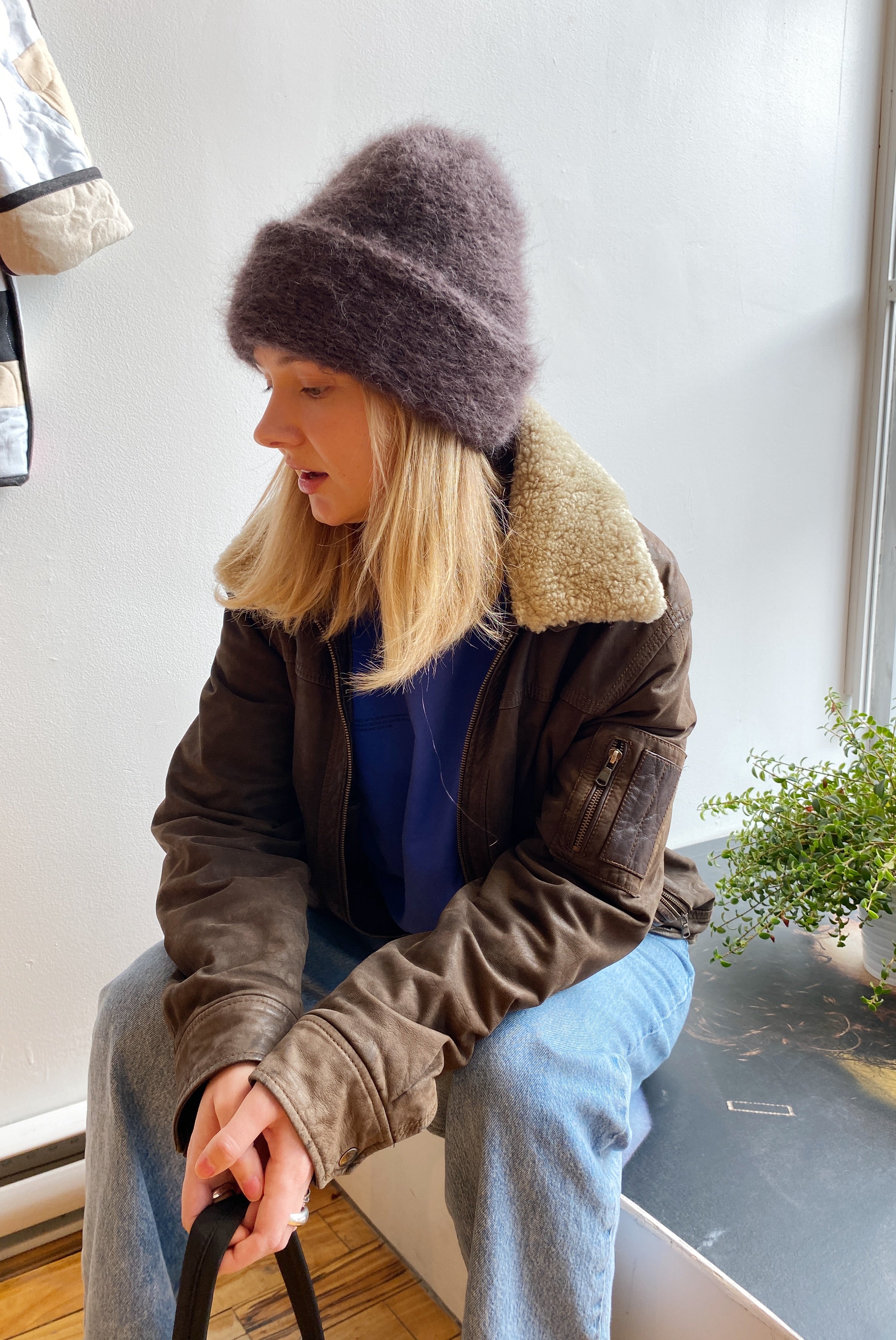 Model with blonde hair and an aviator coat sitting on a ledge, holding a purse, and wearing a fuzzy chocolate brown mohair beanie by Heirloom Hats for a chic, cozy look.