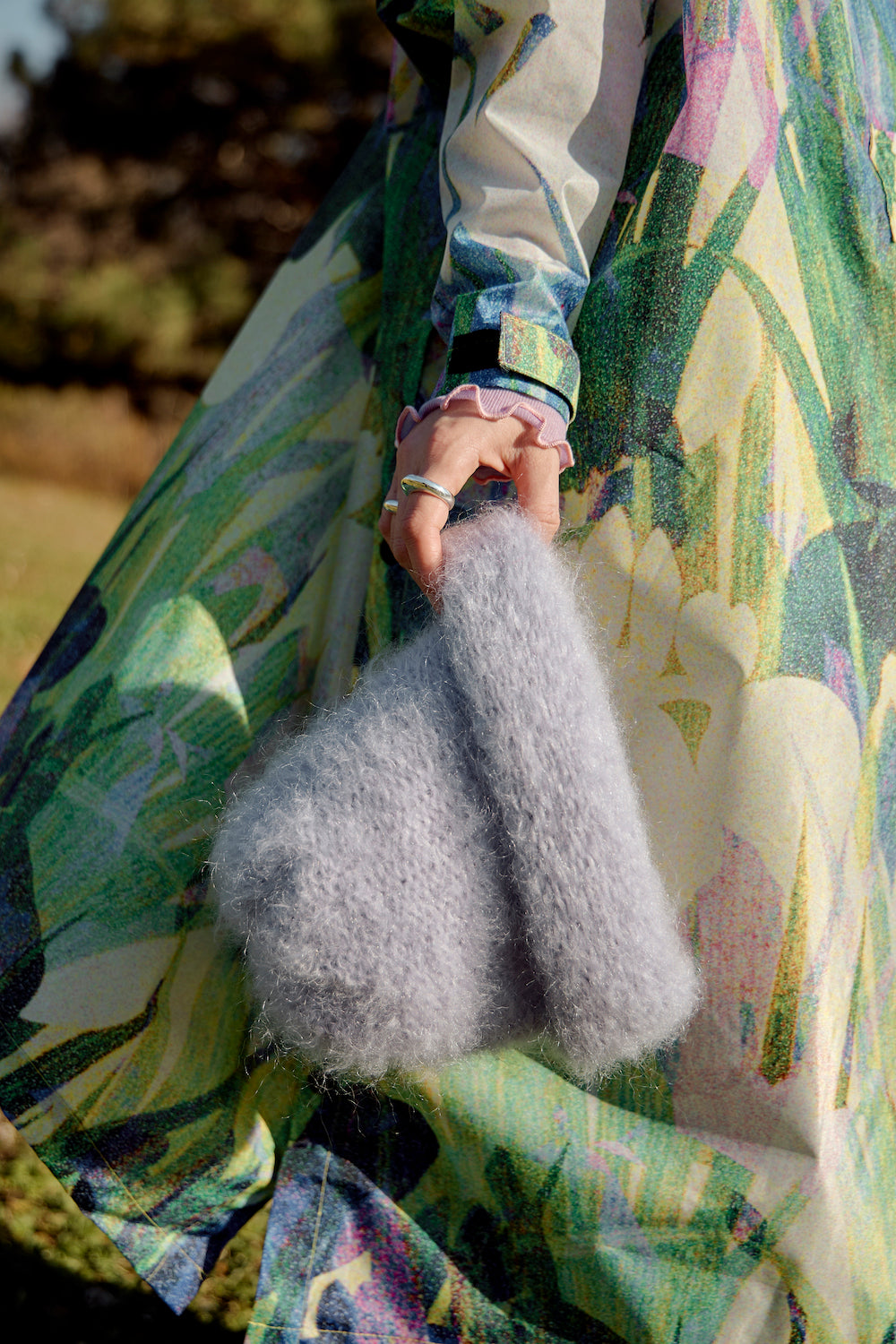 Hand adorned with rings holding a lilac mohair beanie against the fabric of a long raincoat, showcasing a stylish, cozy accessory.