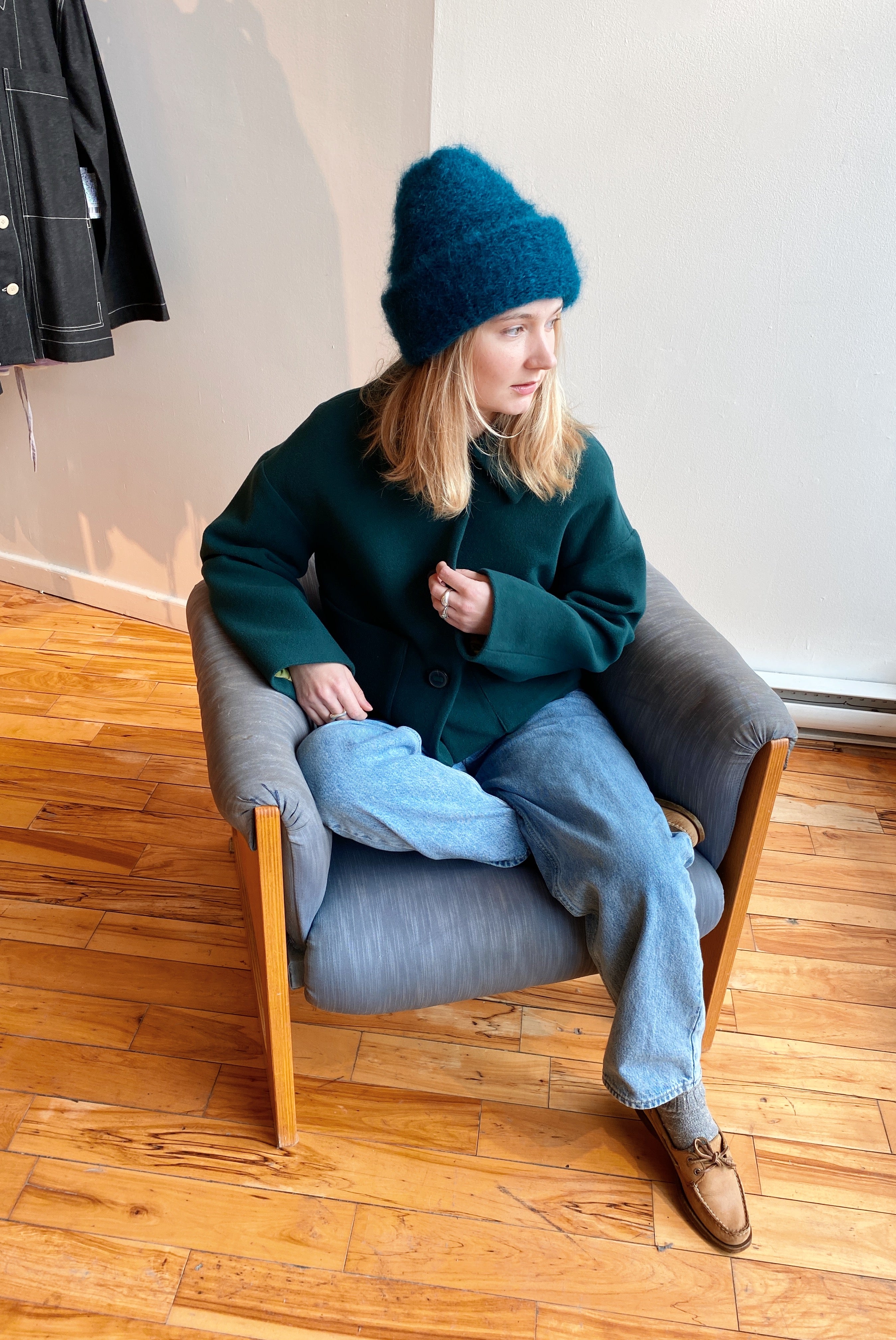 A blond-haired model sits on a mid-century chair, wearing a fuzzy green mohair beanie by Heirloom Hats.