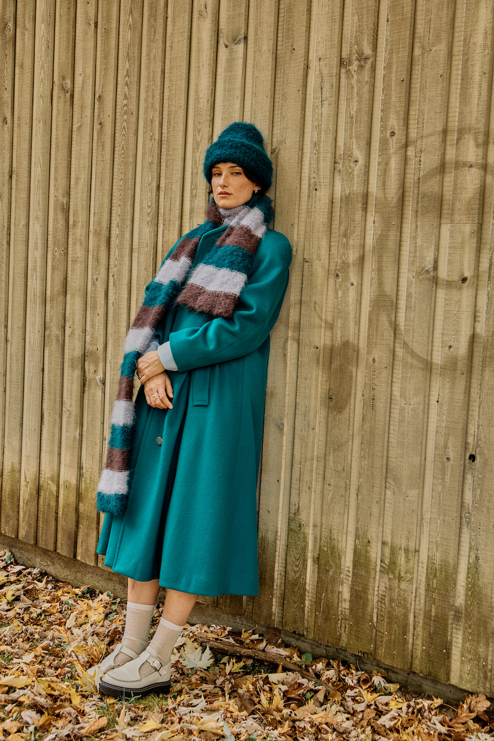 A model with a short brown bob wears a green mohair beanie and striped scarf by Heirloom Hats, leaning against a cabin in the forest.
