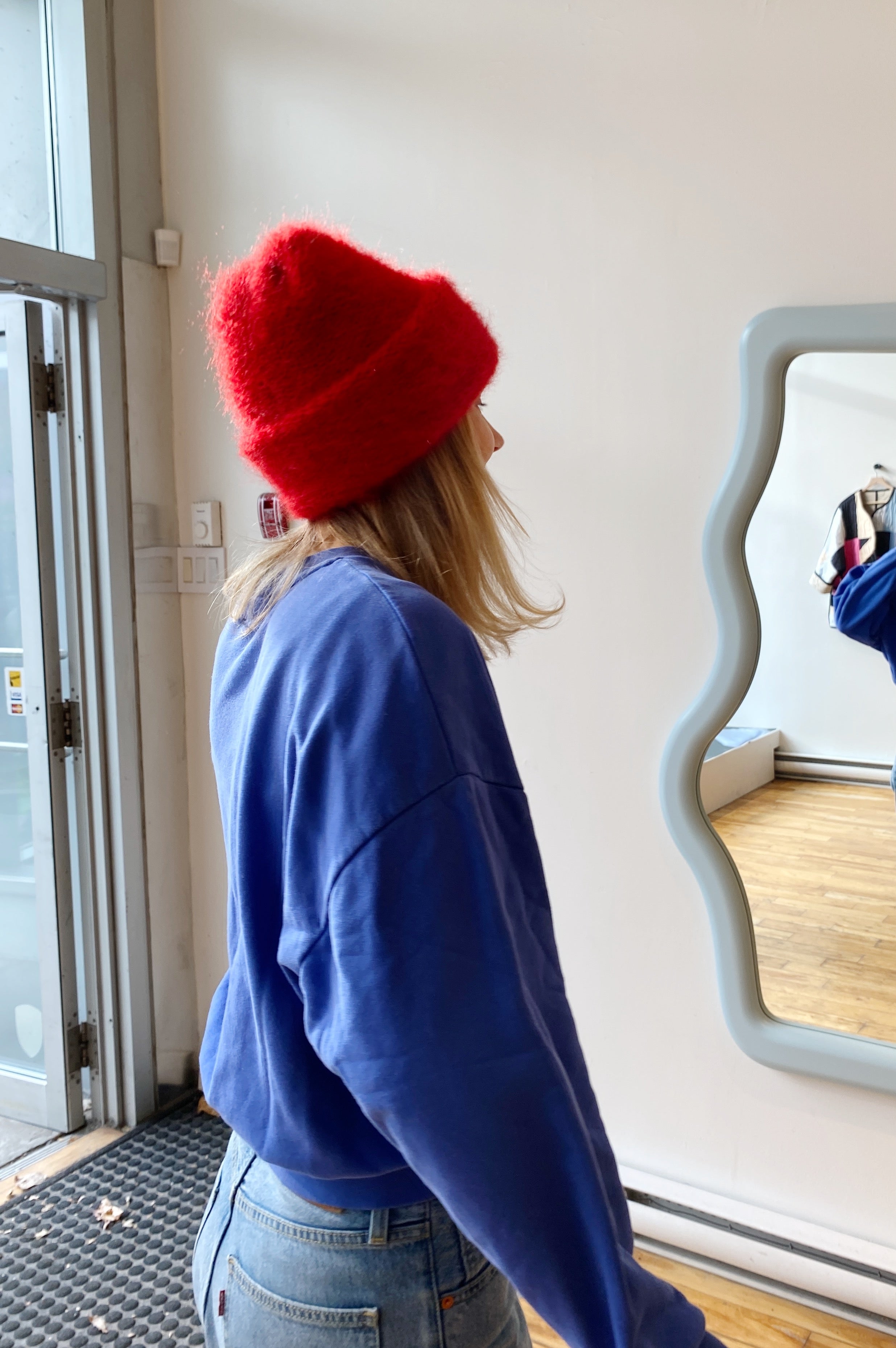 A blond-haired model in a royal blue sweater wears a ruby red mohair beanie by Heirloom Hats, gazing at her reflection in a wavy baby blue mirror.