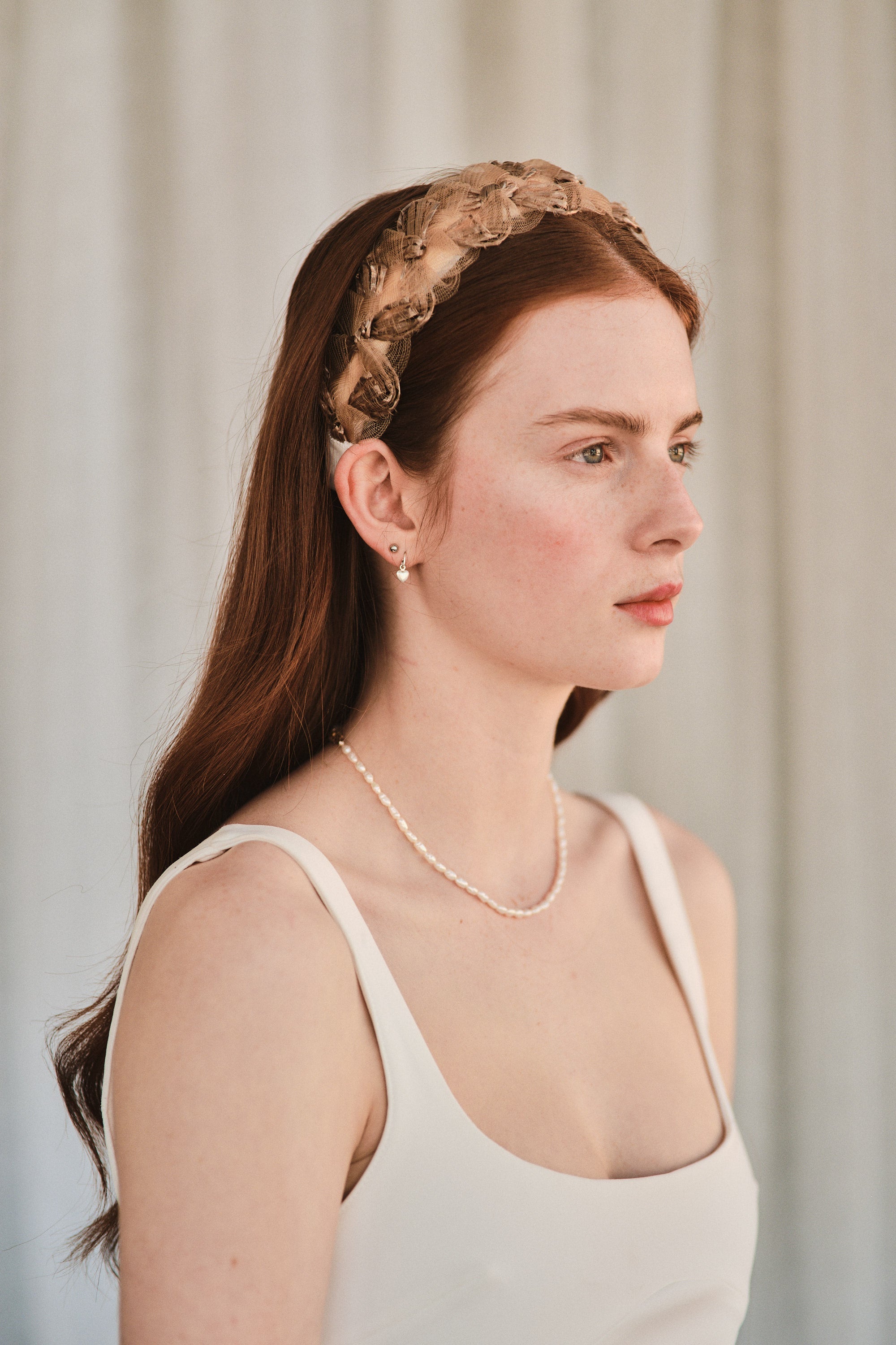 Model with long red hair wears a bronze coloured headband made with deadstock straw, styled with a white evening dress and pearl necklace.