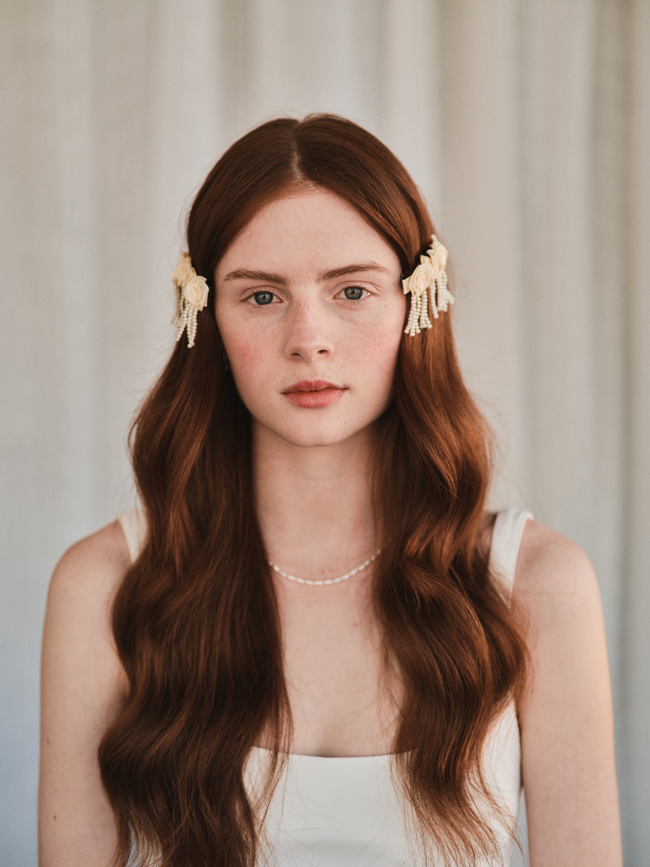 Model with natural red hair wears an off-white satin rosette hair clip with pearl strands, holding hair to the sides of her face.