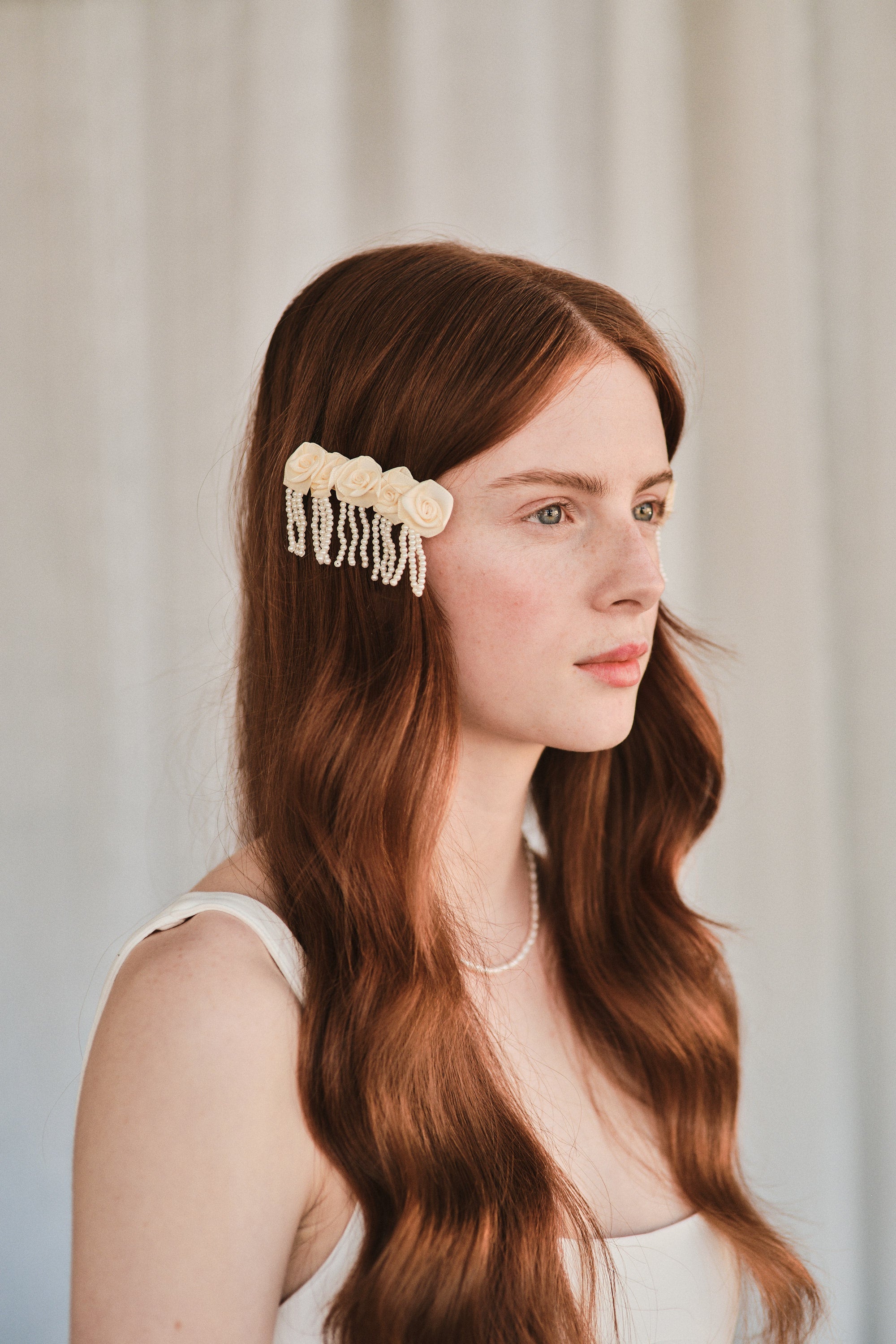 Model with natural red hair wears an off-white satin rosette hair clip with pearl strands, holding hair to the sides of her face.