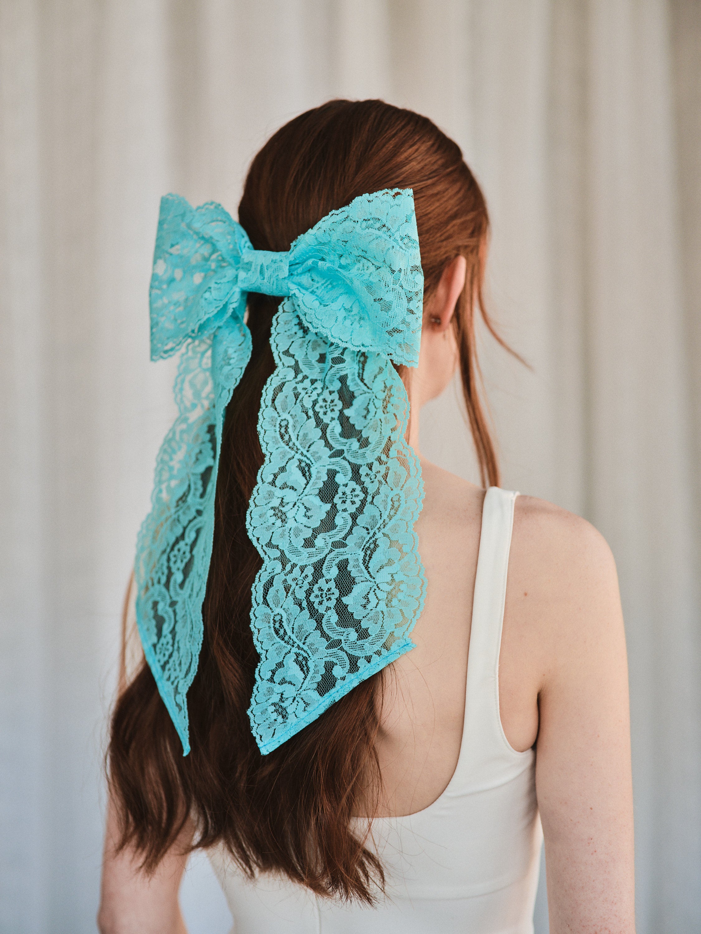 Model with long natural red hair wears an oversized aqua blue lace bow hair clip, styled in a half-up look with a white tank top.