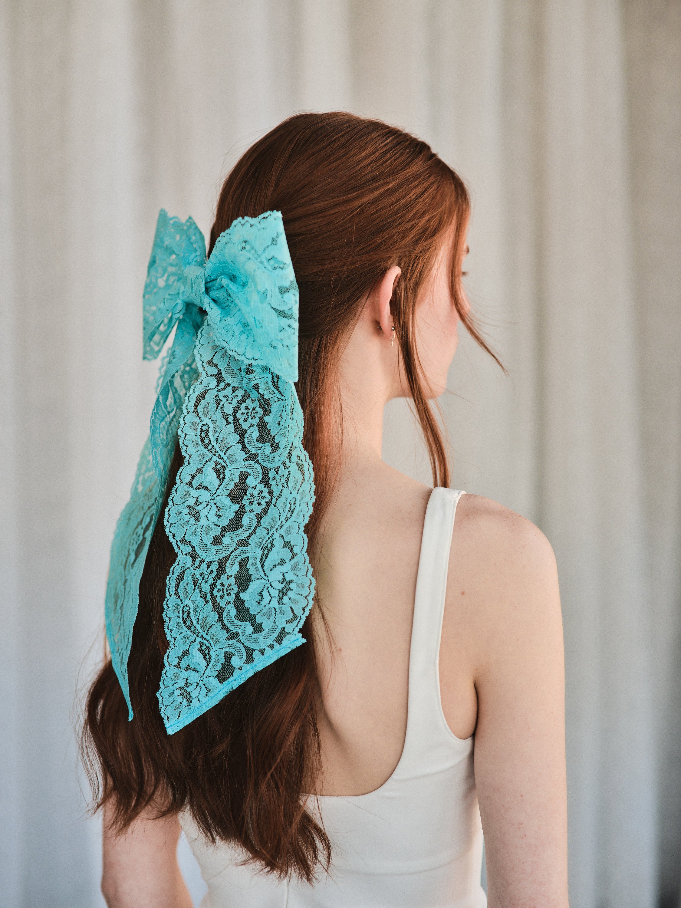 Model with long natural red hair wears an oversized aqua blue lace bow hair clip, styled in a half-up look with a white tank top.