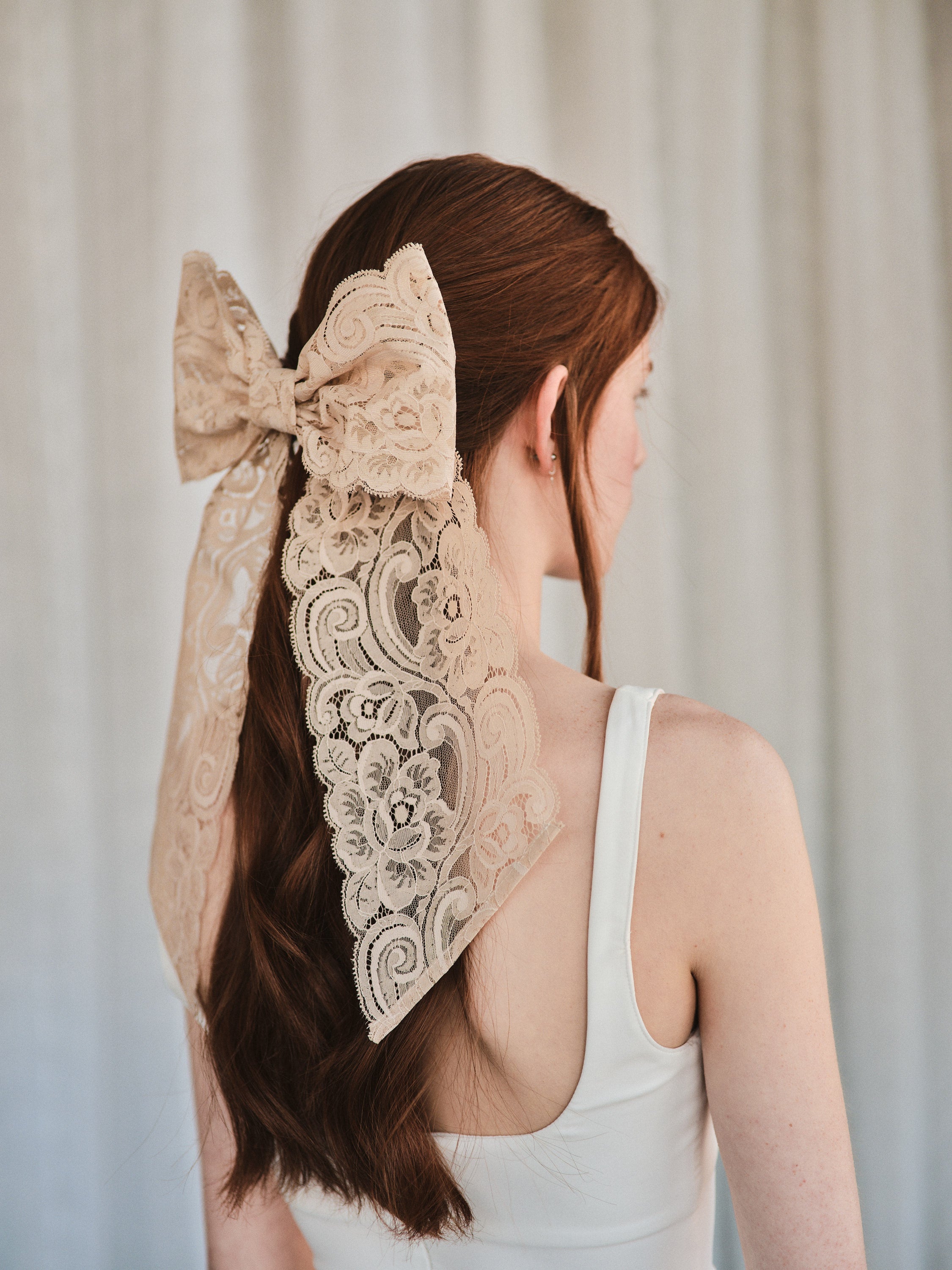 Model with natural red hair wears an oversized champagne coloured lace bow hair clip over low ponytail and a white tank top.