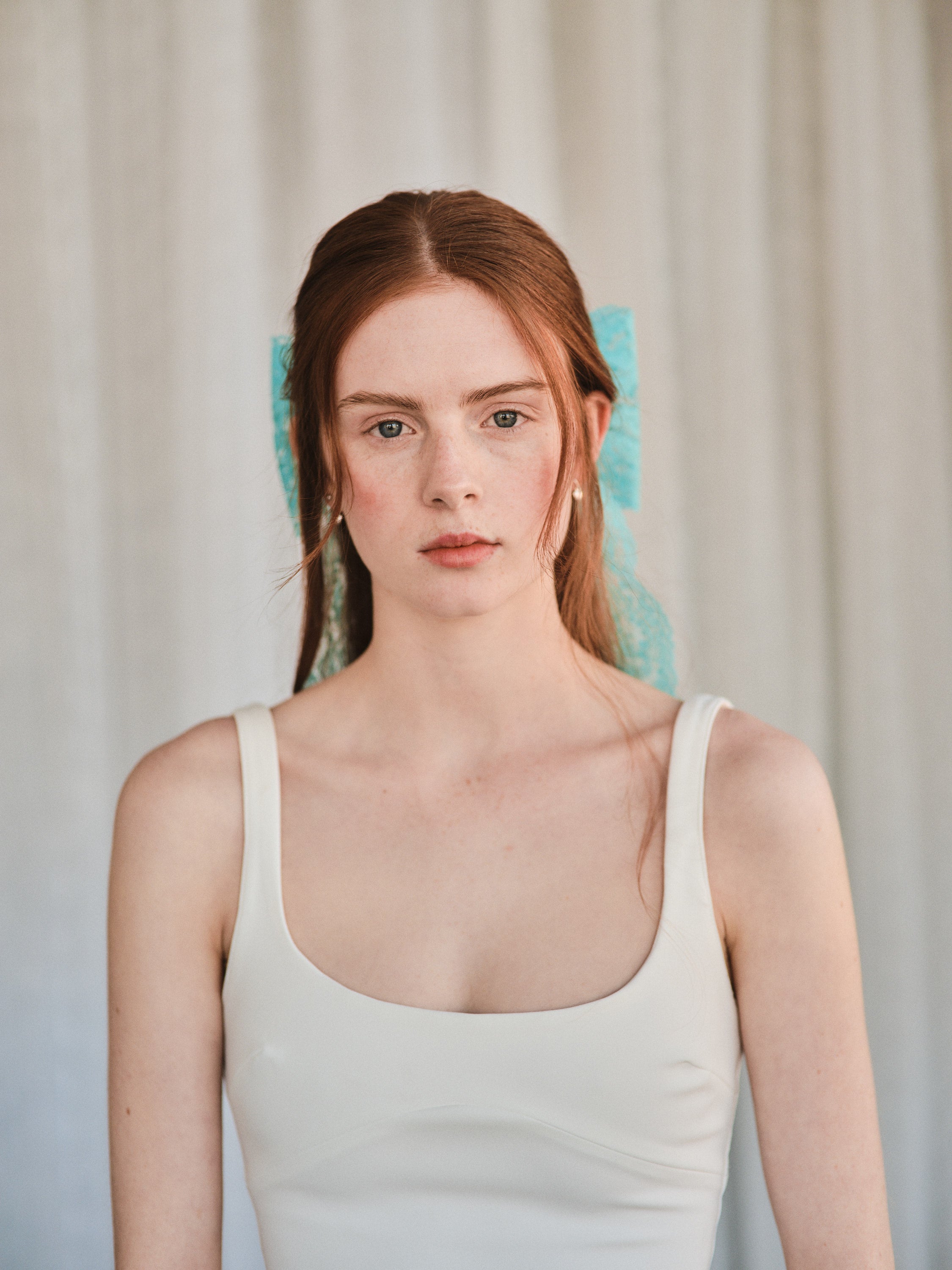 Model with natural red hair wears an oversized aqua blue lace bow hair clip and a white tank top, gazing softly at the camera.