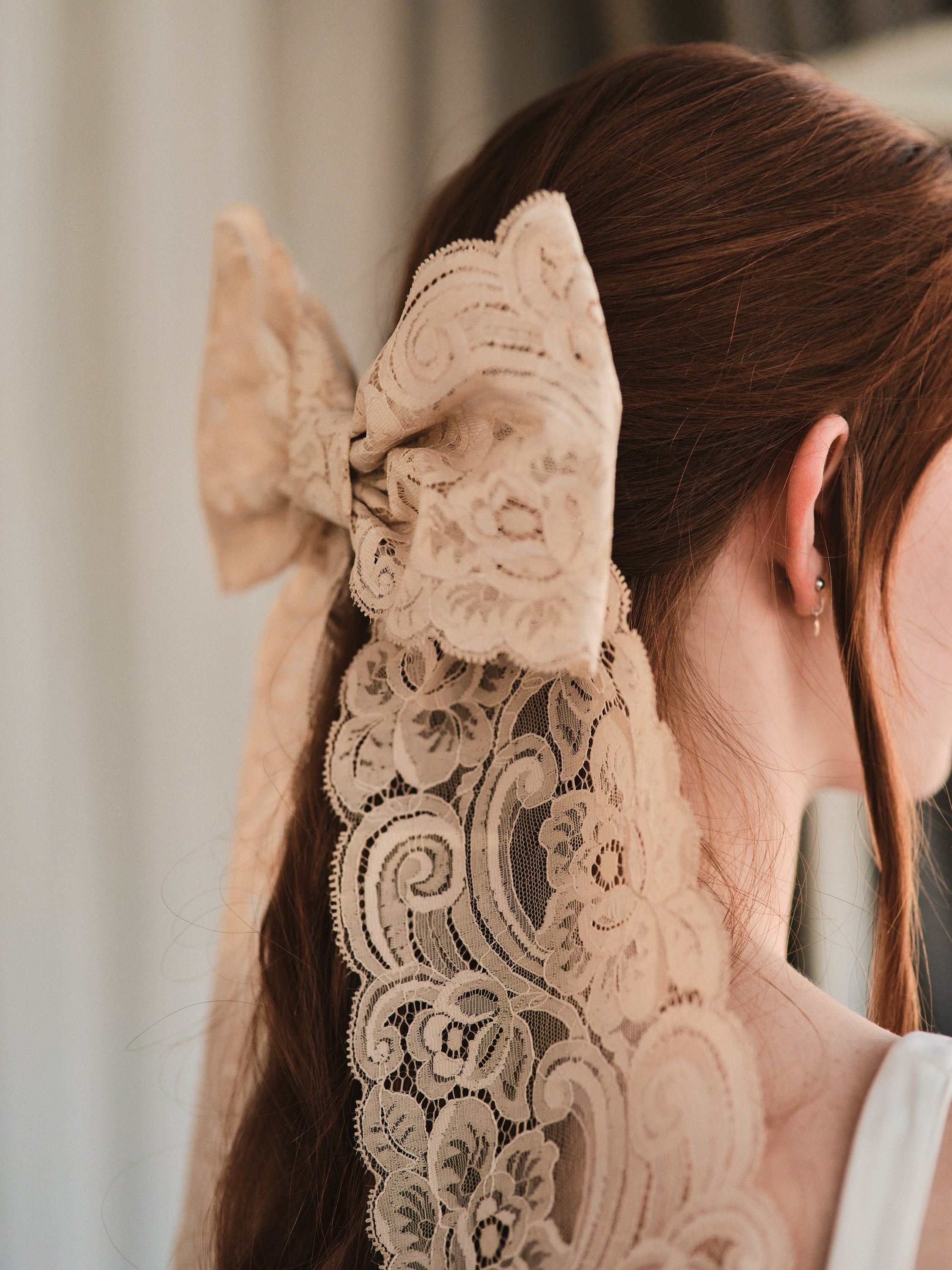 Model with natural red hair wears an oversized champagne coloured lace bow hair clip over low ponytail and a white tank top.