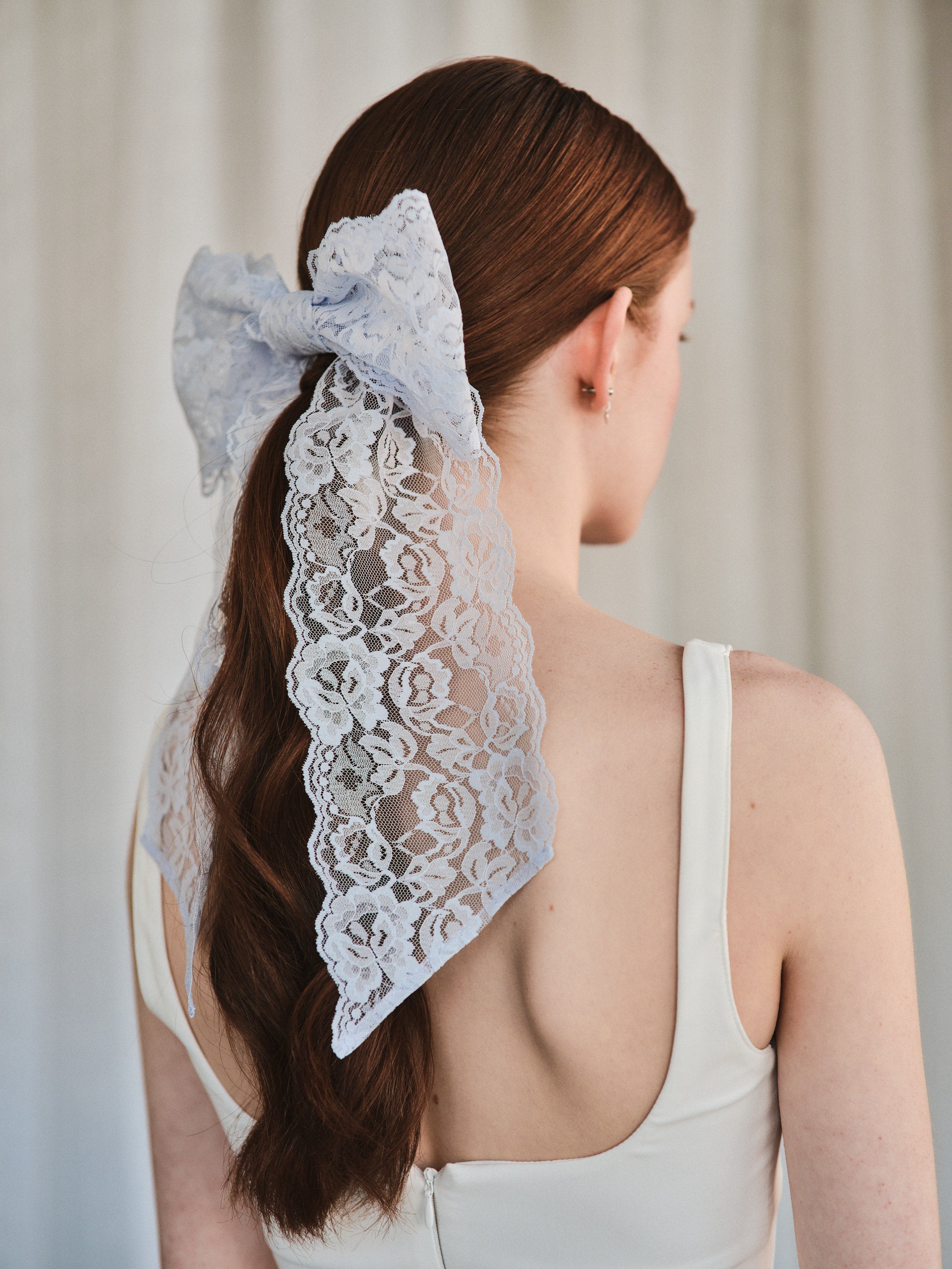 Model with natural red hair wears an oversized light purple lace bow hair clip over low ponytail and a white tank top.