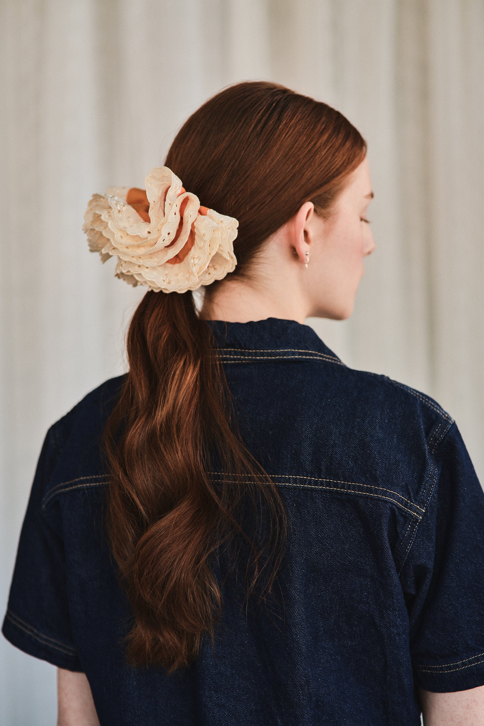 Azalea hair clip featuring guava orange linen and off-white eyelet lace, worn over a ponytail on a woman with natural long red hair. She’s wearing a dark denim shirt and is shown from a back profile with her head to the side.