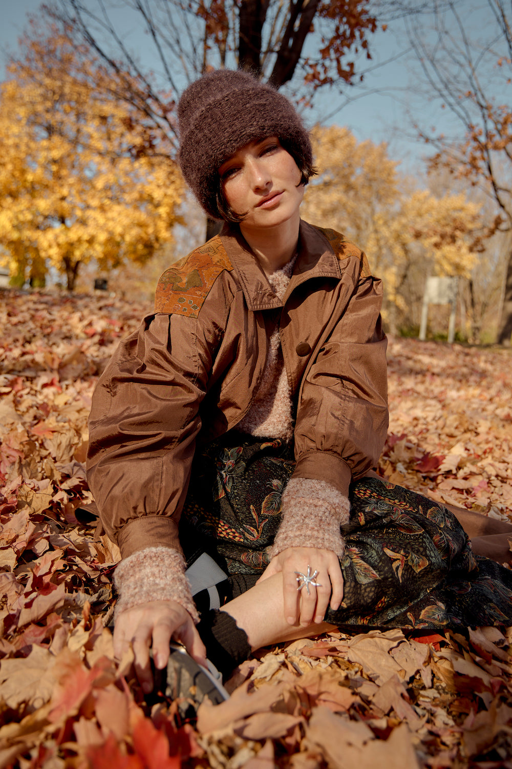 Model wearing a chocolate brown mohair beanie by Heirloom Hats, lying in a sea of fallen leaves, surrounded by vibrant autumn trees on a sunny fall day.