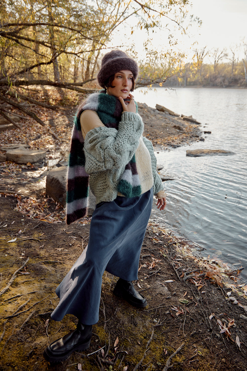 Model wearing a chunky knit sweater, chocolate brown mohair beanie, and striped scarf by Heirloom Hats, standing by a lake surrounded by trees on a warm autumn day.