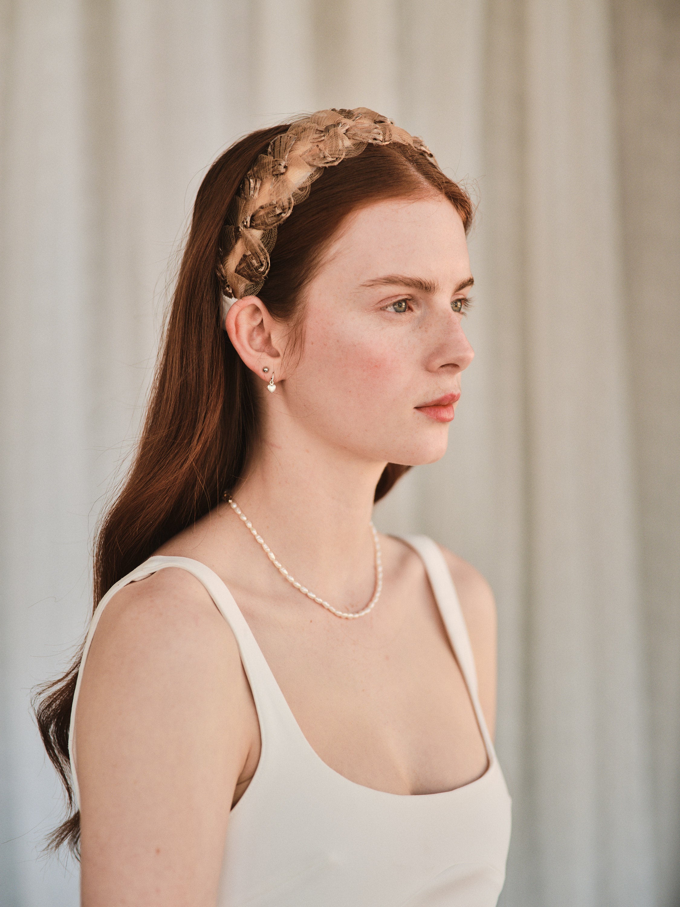 Model with long red hair wears a bronze coloured headband made with deadstock straw, styled with a white evening dress and pearl necklace.
