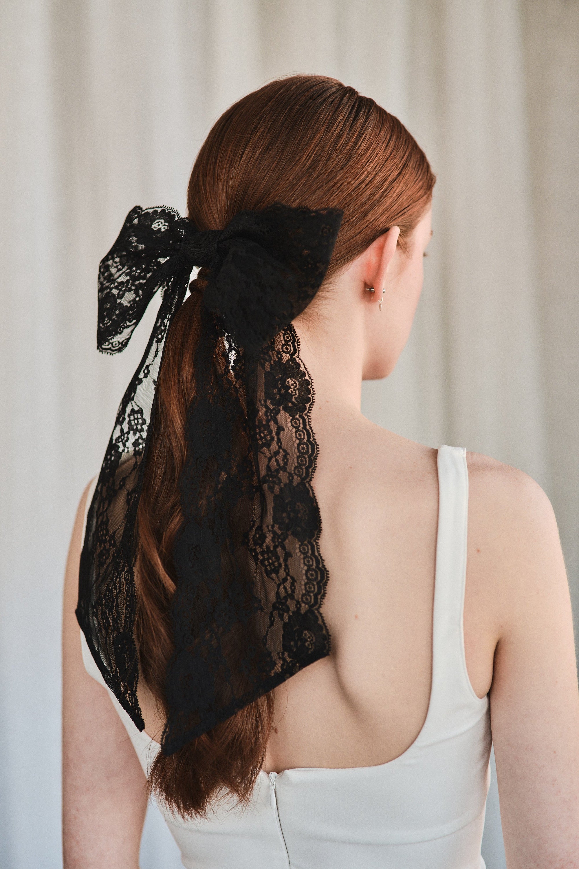 Model with natural red hair wears an oversized black lace bow hair clip styled in a low ponytail and a white tank top.