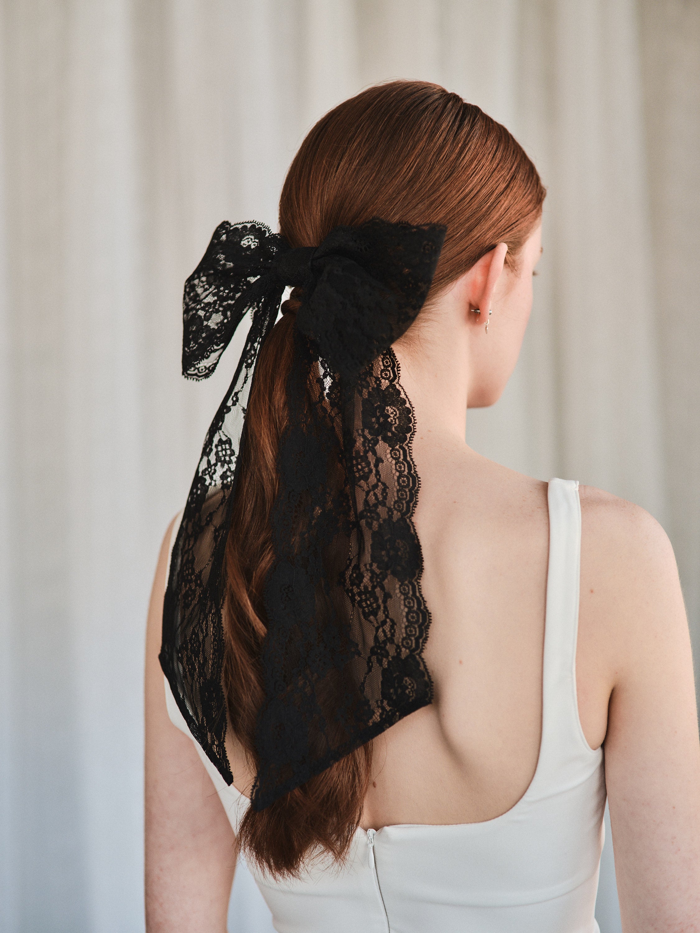 Model with natural red hair wears an oversized black lace bow hair clip styled in a low ponytail and a white tank top.