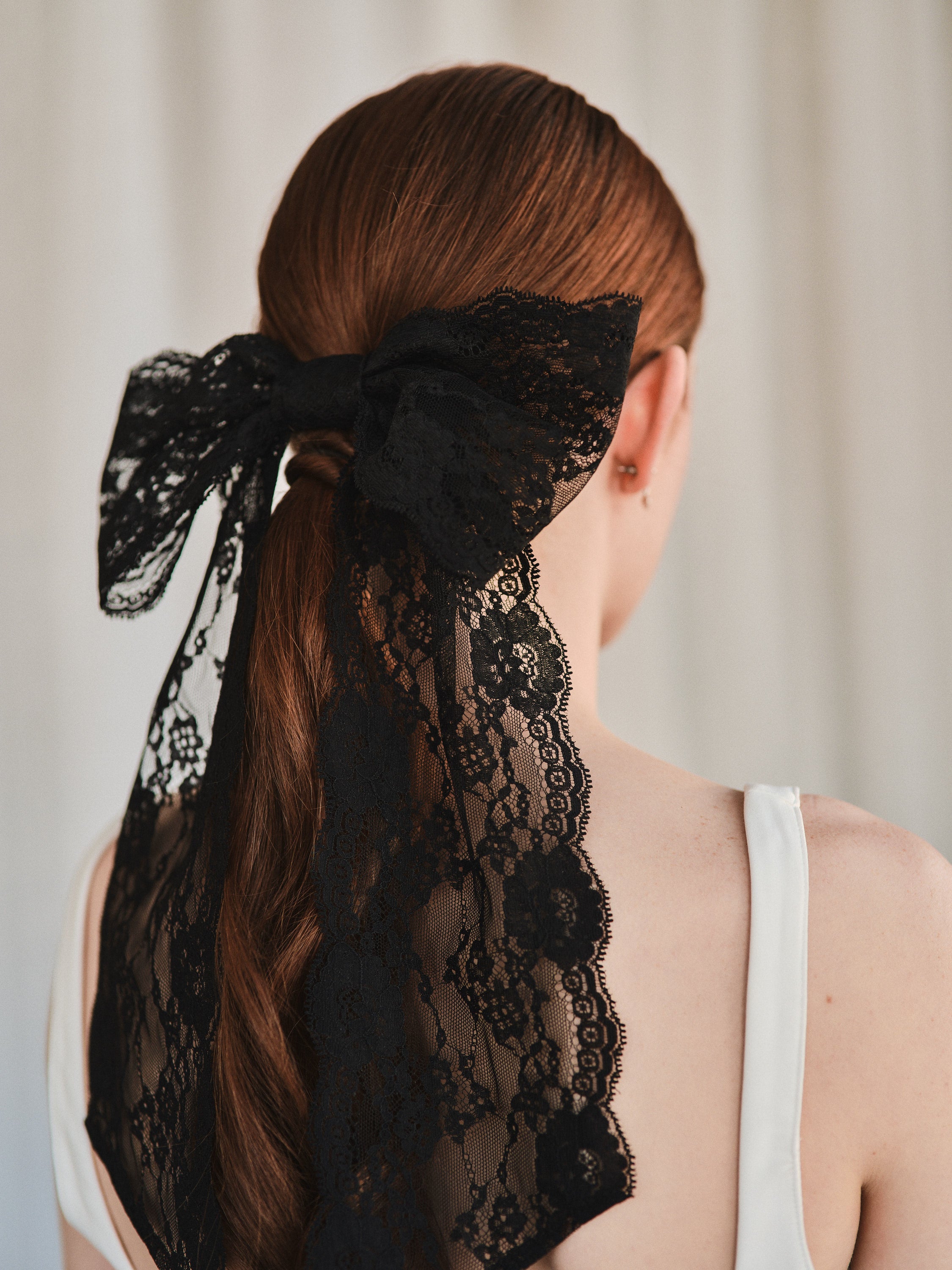 Model with natural red hair wears an oversized black lace bow hair clip styled in a low ponytail and a white tank top.
