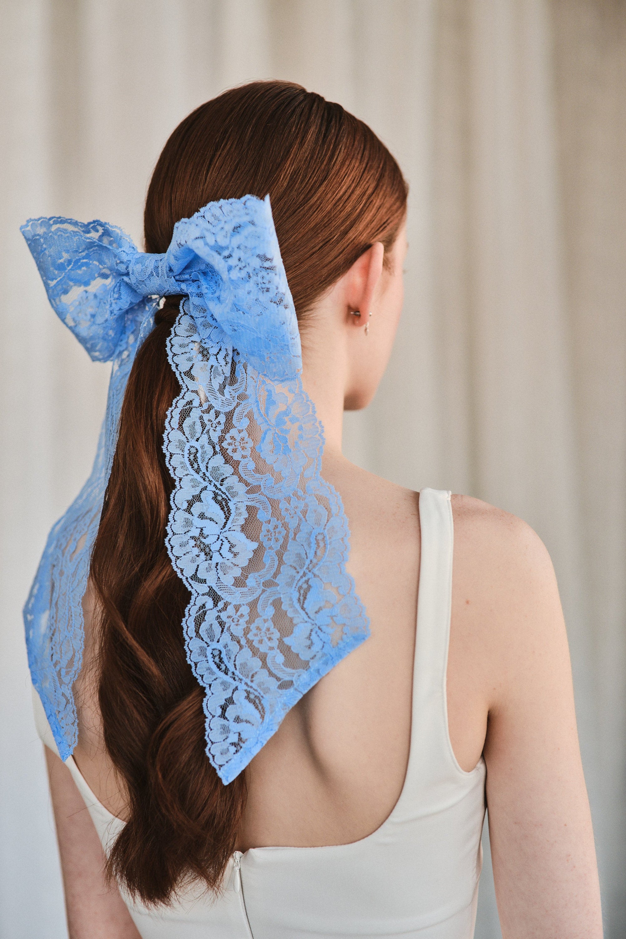 Model with natural red hair wears an oversized cornflower blue lace bow hair clip over low ponytail and a white tank top.