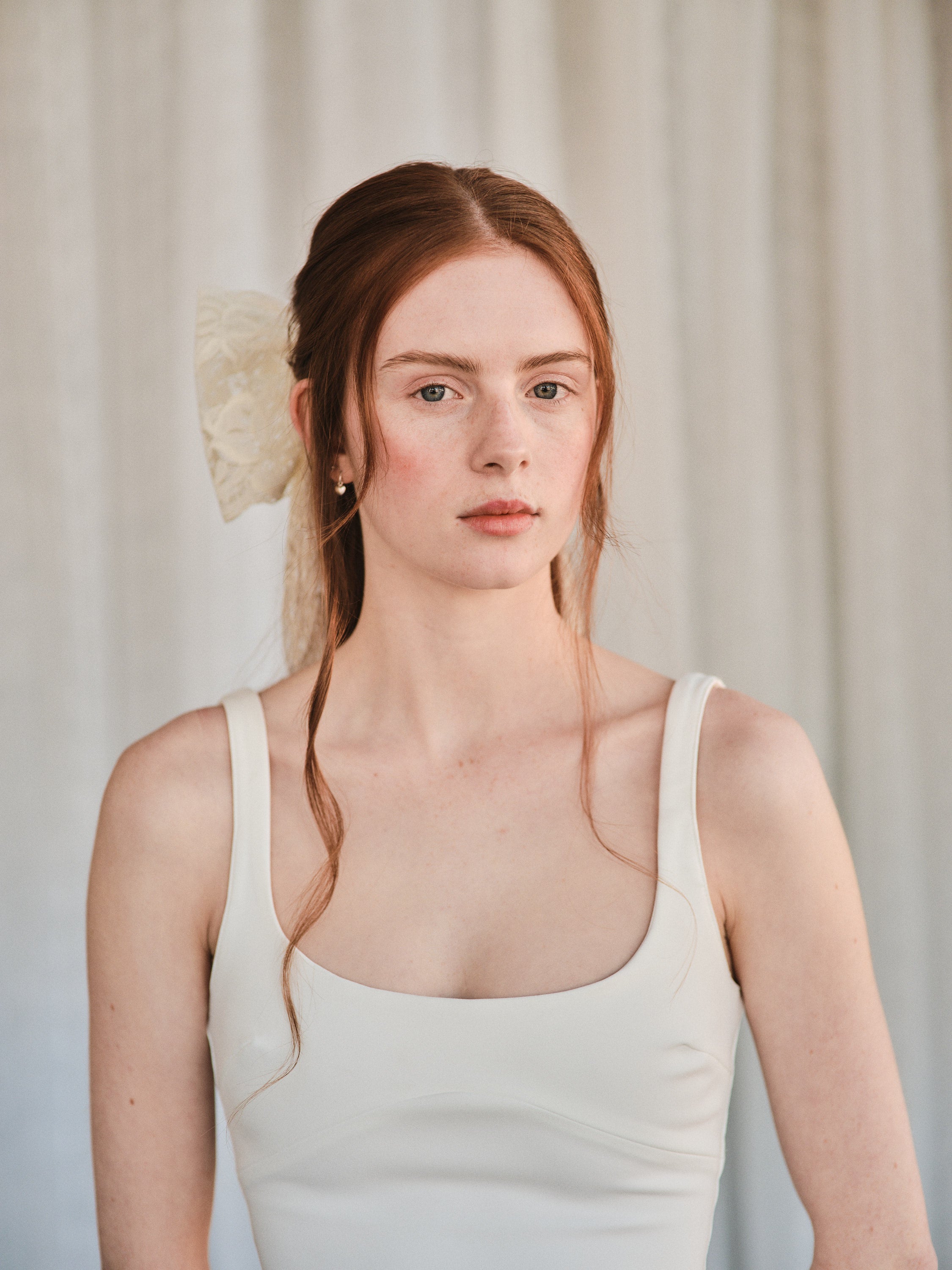 Model with natural red hair wears an oversized off white lace bow hair clip styled in a half-up look and a white tank top.