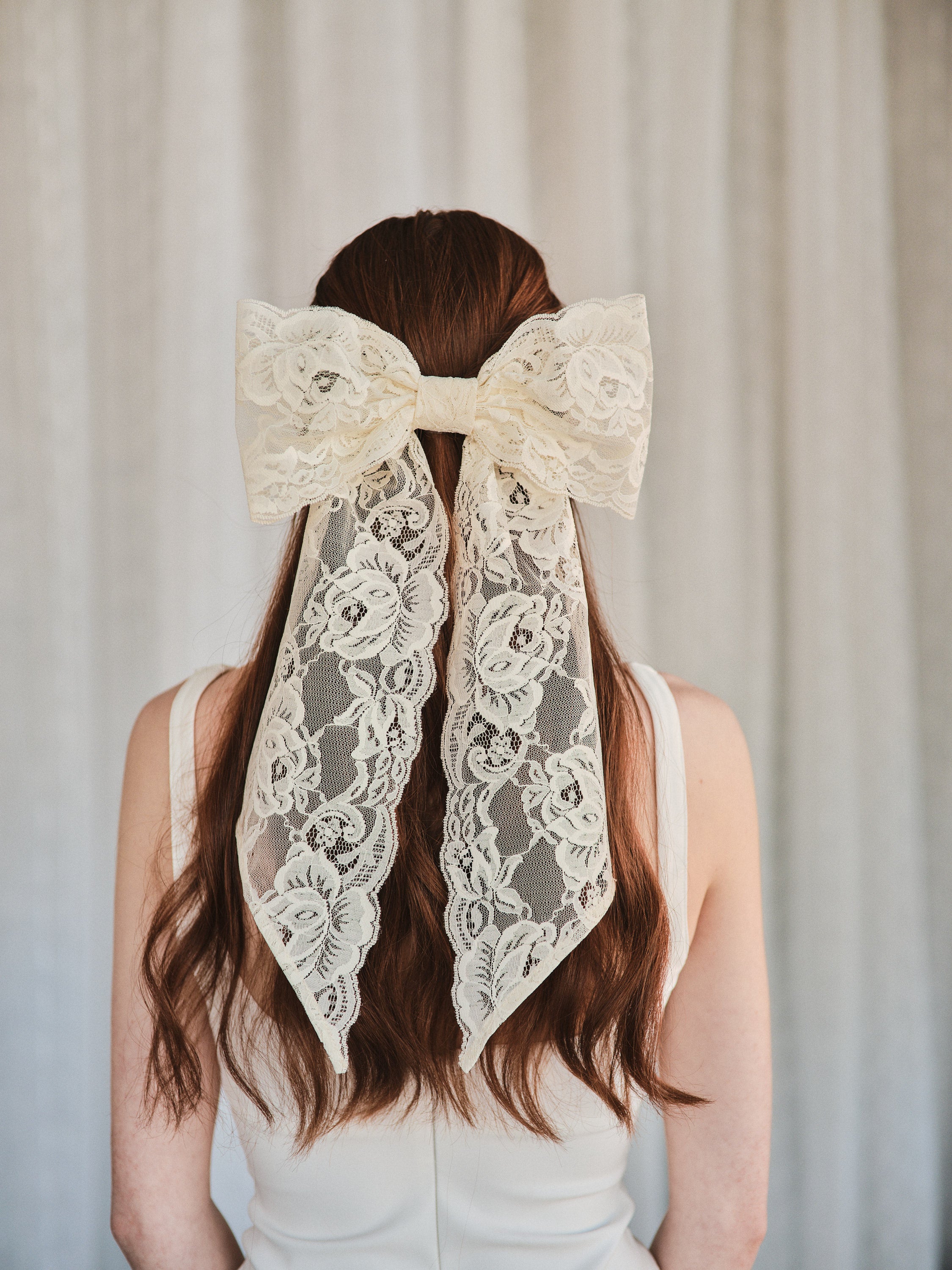 Model with natural red hair wears an oversized off white lace bow hair clip styled in a half-up look and a white tank top.