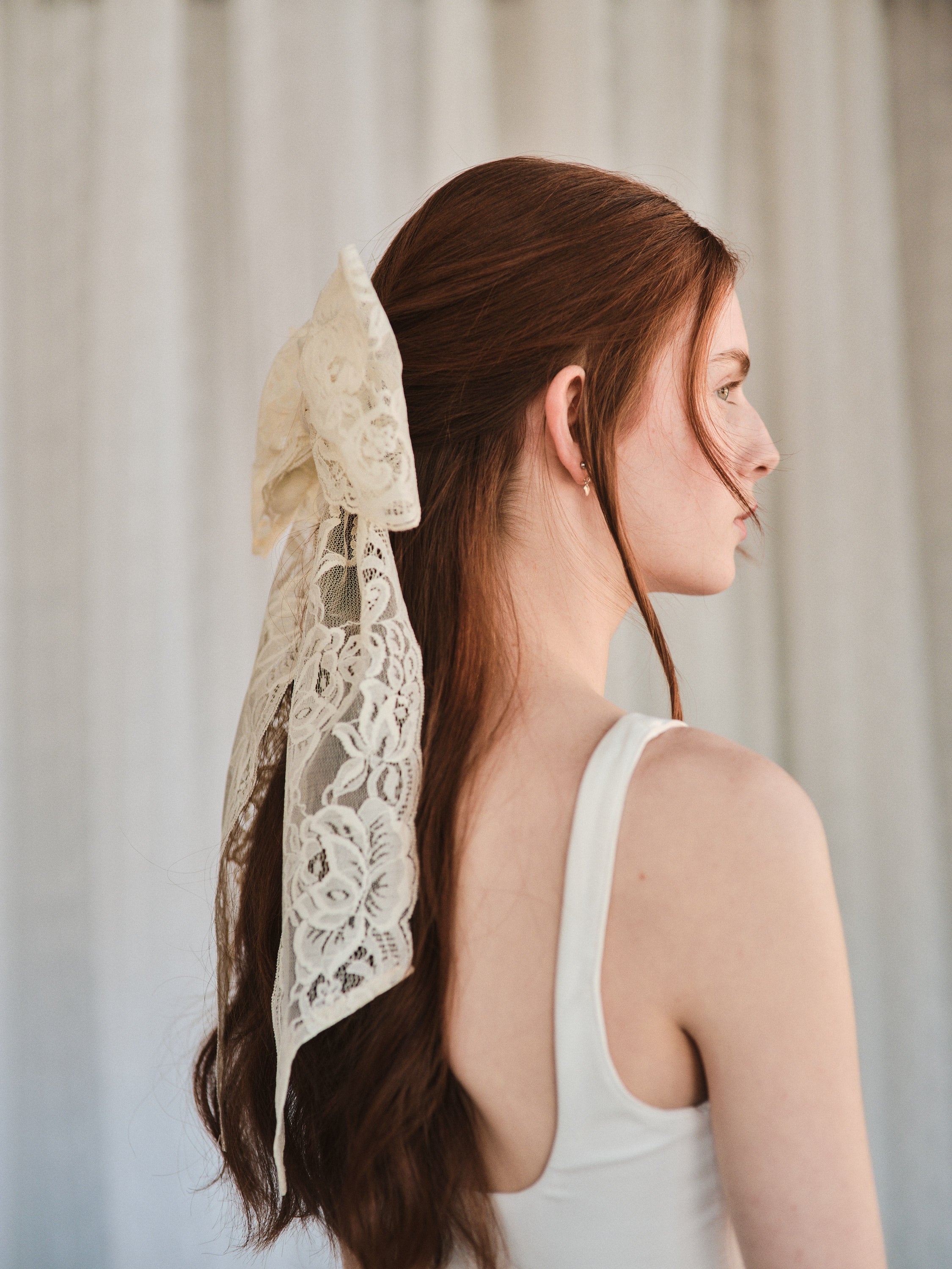 Model with natural red hair wears an oversized off white lace bow hair clip styled in a half-up look and a white tank top.