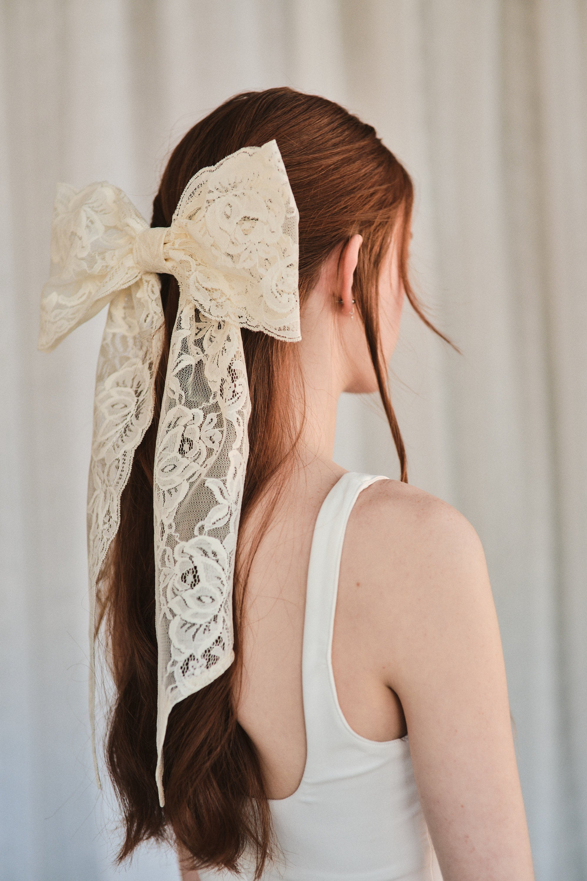 Model with natural red hair wears an oversized off white lace bow hair clip styled in a half-up look and a white tank top.
