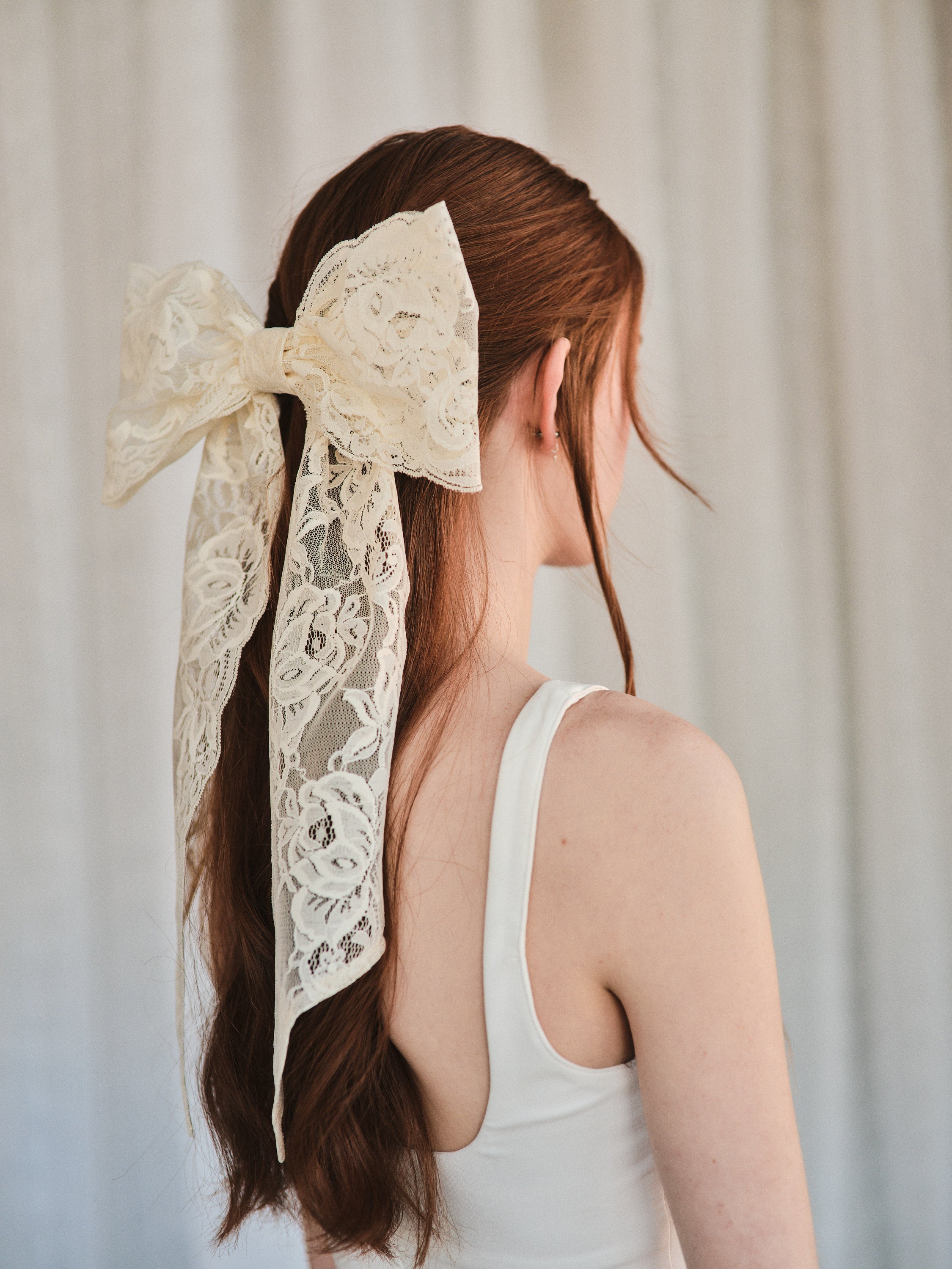Model with natural red hair wears an oversized off white lace bow hair clip styled in a half-up look and a white tank top.