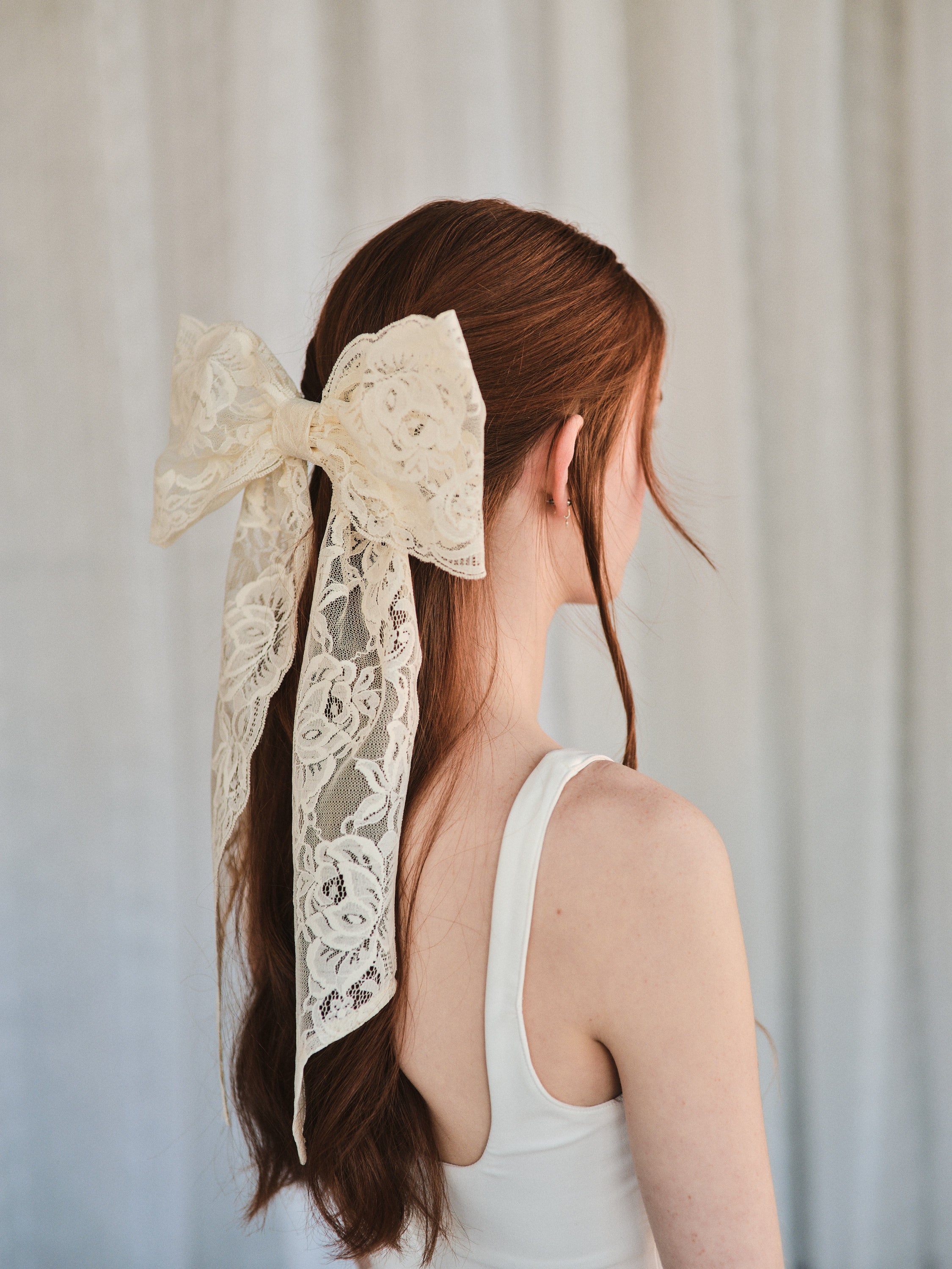 Model with natural red hair wears an oversized off white lace bow hair clip styled in a half-up look and a white wedding dress.